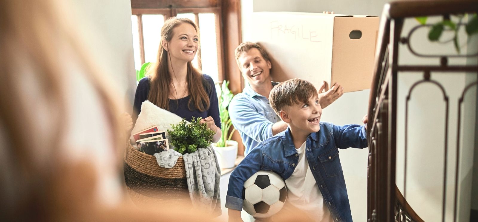 Young boy with soccer ball, mother with basket, and father with large cardboard box walking up staircase as they move into new home